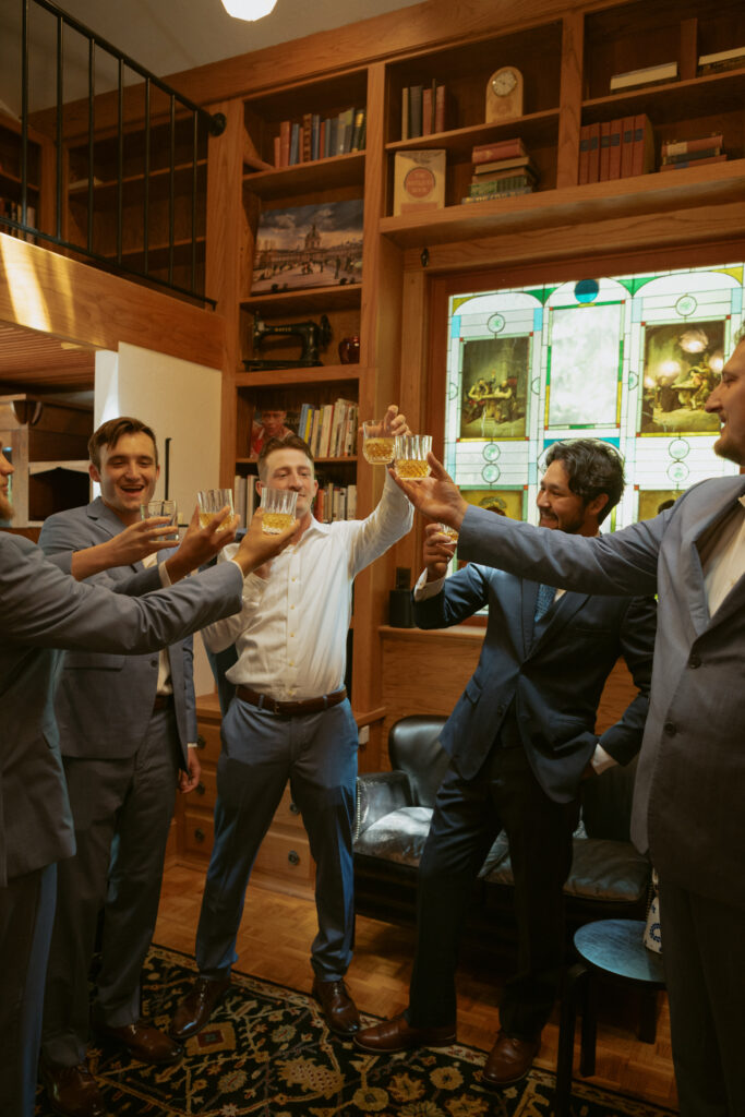 Groom and groomsmen getting ready together, adjusting ties and jackets in a candid wedding morning moment.